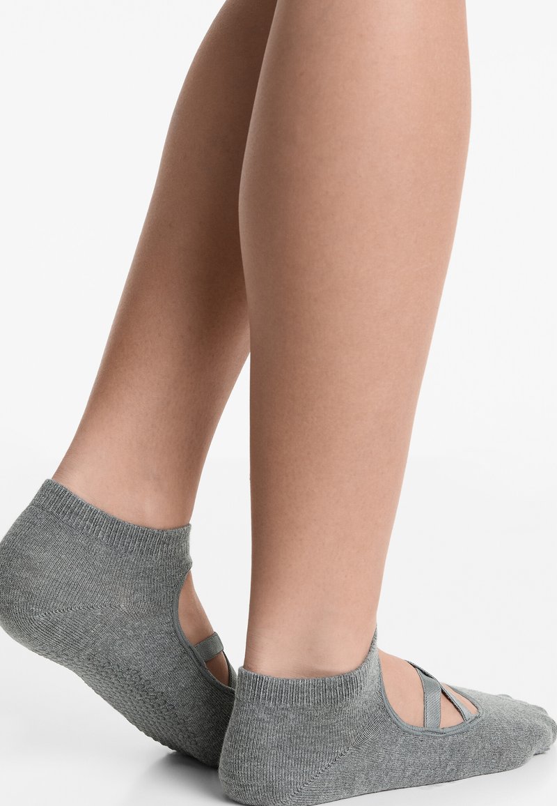 Pieds portant des chaussettes grises à la cheville avec des bandes élastiques croisées, vus de côté et du talon sur fond blanc.