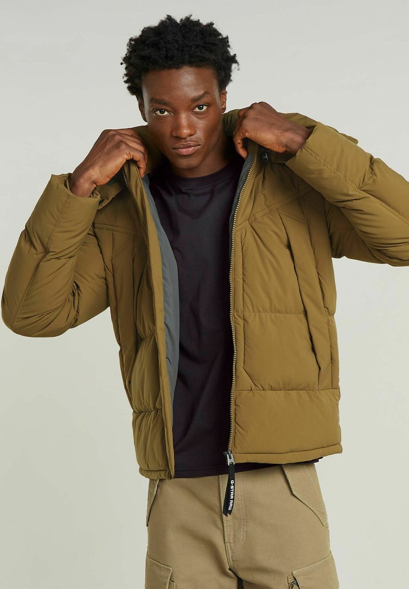 Oliwkowa kurtka puchowa z zamkiem z przodu, z szarym wykończeniem, poziomym pikowaniem i bocznymi kieszeniami. Noszona z czarną koszulą.