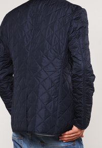 Marinefarbene gesteppte Jacke mit tailliertem Design, die ein Rautenmuster und strukturierte Schultern aufweist. Zeigt die Rückansicht mit strukturiertem Stoff und Nahtdetails.