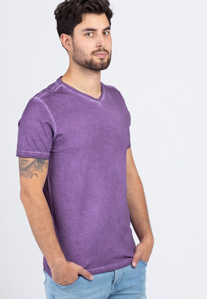 Purple cotton t-shirt with a v-neck, short sleeves, and a slightly textured finish. Model poses with hands in pockets, wearing light blue jeans.