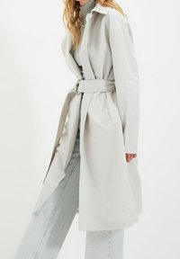 Mujer vistiendo un abrigo trench ligero de color gris con cinturón, sobre un suéter de cuello alto gris y pantalones vaqueros de pierna ancha, de pie frente a un fondo blanco.