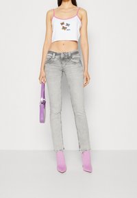 Woman wearing white crop top with butterfly print, gray jeans, pink patterned boots, holding small purple handbag against white background.