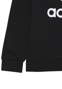 Černá mikina s ribkovanou strukturou. Na přední straně se nachází bílé logo "adidas". Manžety a lem jsou také ribkované.