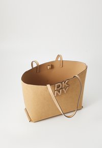 Beige tote bag med en strukturert form, laget av mykt materiale. Har et logo-accent, to håndtak og et minimalistisk design.