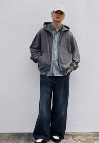Übergroßer grauer Hoodie mit Fronttaschen, hellblaue Knopfleiste darunter, dunkle weit geschnittene Jeans und schwarz-weiße Schuhe.