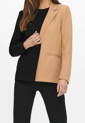 Femme portant un blazer bicolore, noir à gauche et beige à droite, associé à un pantalon noir et un haut noir.