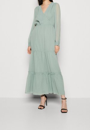 Pitkä mintunvihreä mekko, jossa on pilkkuja, V-aukko, läpinäkyvät hihat ja kerroksittainen hame. Yhdistetty mustien remmikenkien kanssa.