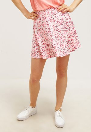 Vrouw draagt een roze A-lijn rok met bloemenpatroon, koraalkleurige top en witte sneakers met veters, staand met handen in de zij tegen een effen achtergrond.