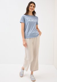 Vaaleansininen kuvioitu t-paita lyhyillä hihoilla, jossa on graafinen printti. Yhdistetty vaaleanbeigeihin leveisiin housuihin ja vaaleansinisiin lenkkareihin.