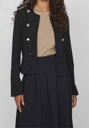 Woman wearing a black cropped blazer with silver buttons over a beige top and a high-waisted black pleated skirt.