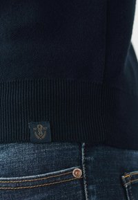 Pulover tricotat bleumarin cu manșetă côtée, având un mic etichet cu un logo și anul "1982". Asortat cu blugi din denim închis, evidențiind detalii de cusătură.