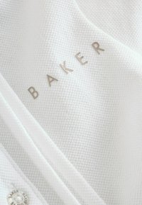 Valkoinen, teksturoitu kangas, jossa on hillitty "BAKER"-logo metallikirjaimin, ja alla nappi, jossa on sama merkkitunnus.