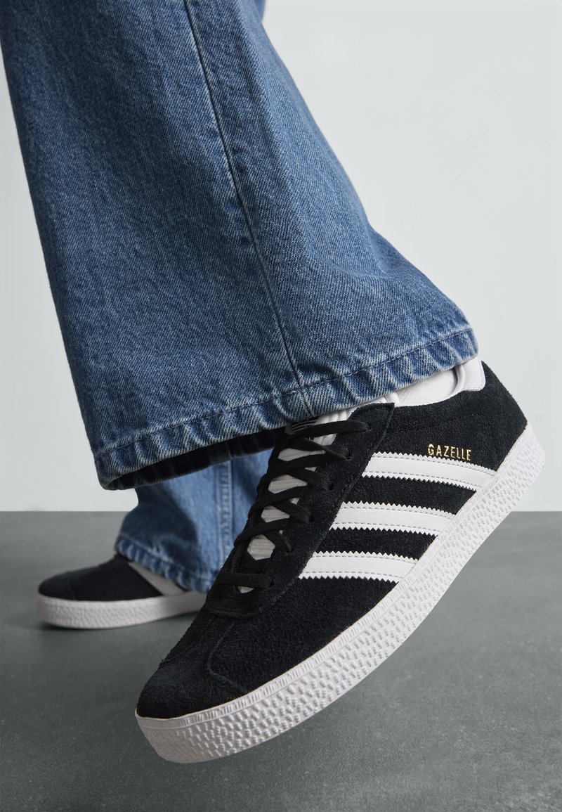 Personne portant un jean bleu et des baskets Adidas Gazelle noires avec des rayures blanches, debout sur un sol gris.