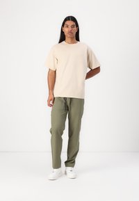 Beige katoenen t-shirt met korte mouwen, gecombineerd met groene cargo pants met zijzakken, en witte sneakers op een model tegen een effen achtergrond.