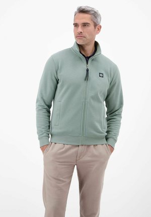 Mann mit grauen Haaren, der eine hellgrüne Jacke mit Reißverschluss und beigefarbene Hose trägt, steht mit den Händen in den Taschen und blickt nach links vor weißem Hintergrund.