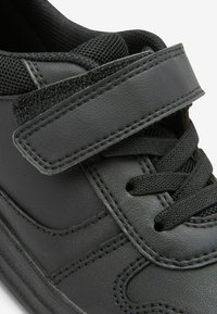Baskets synthétiques noires avec un col rembourré, une languette en maille, des lacets et une sangle Velcro. Surfaces texturées et détails de coutures contrastées.