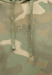 Tissu à motif camouflage avec logo "GAP" brodé et deux cordons verts d'une capuche.