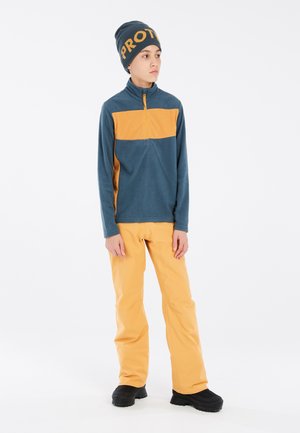 Pull en polaire bleu avec une bande orange, associé à un pantalon orange et des bottes noires. Le bonnet arbore un texte en orange en caractères gras "PROT".