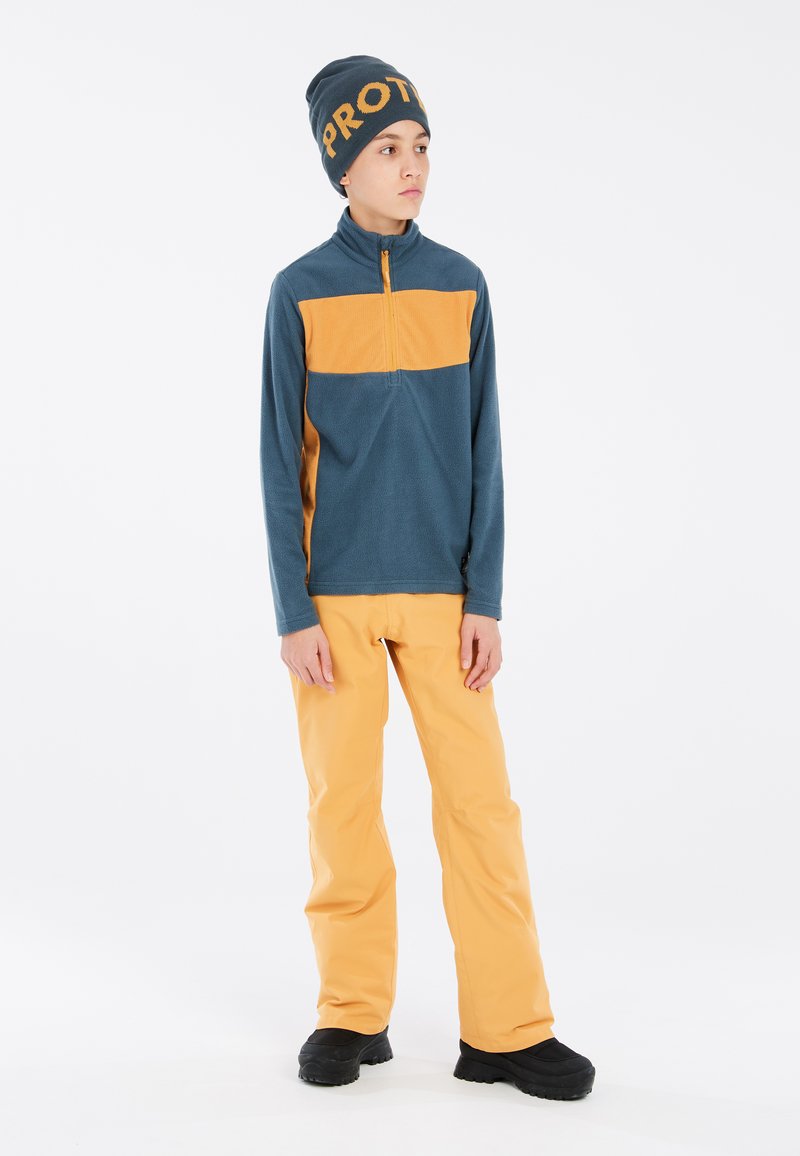 Fleece pullover i blå med orange stribe, parret med orange bukser og sorte støvler. Beanieen har fed "PROT" tekst i orange.