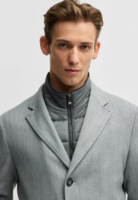 Grå kavaj med texturerad tyg, med en zippered, vadderad grå innerfoder. Kavajen har en enkel knapp och snittlappar.