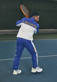 Karaliski zils un balts sporta uzvalks ar "LACOSTE" logotipu dzeltenā krāsā, apvienots ar baltām un zaļām tenisa kurpēm, kuras valkā cilvēks kortā.