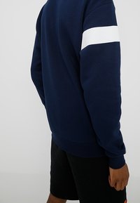 Mörkblå sweatshirts med ett vitt horisontellt rand på vänster ärm. Tillverkad av ett mjukt tyg, med ribbad avslutning på ärmar och nederkant.