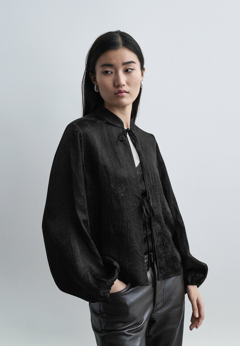 Γυναίκα με μακριά σκουρόχρωμα μαλλιά, που φοράει μια μαύρη μπλούζα με υφή και φουσκωτά μανίκια και μαύρο δερμάτινο παντελόνι, στέκεται με το ένα χέρι στην τσέπη.