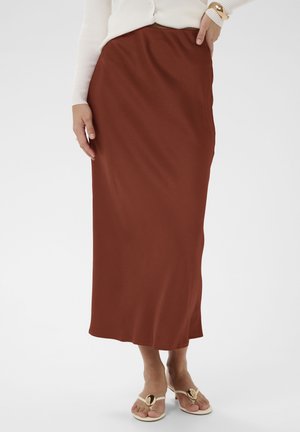 Woman wearing a rust-colored midi skirt, white long-sleeve top, gold watch, and cream sandals with gold accents, standing on a white background.