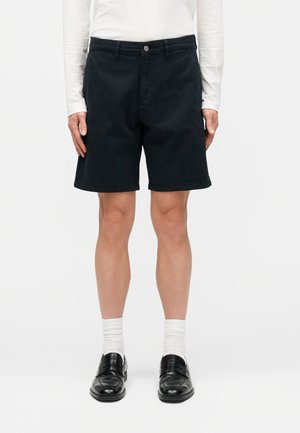 Personne portant un short noir jusqu'aux genoux, un haut blanc à manches longues, des chaussettes blanches et des chaussures habillées noires, debout devant un fond uni.