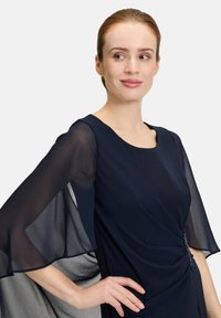 Granatowa sukienka z przezroczystymi, falbaniastymi rękawami, okrągłym dekoltem, skręconym przodem i dekoracyjnym akcentem z metalowymi elementami w talii. Gładka faktura.