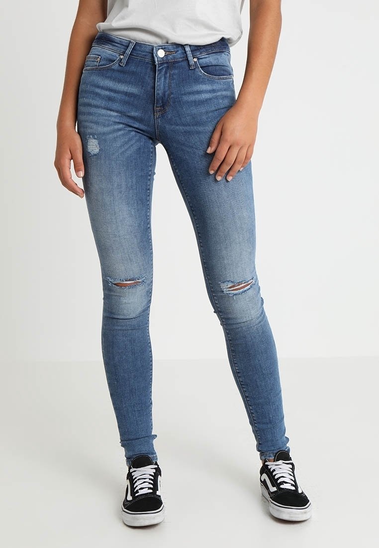 Femme portant un jean skinny bleu taille mi-haute avec des déchirures aux genoux, un t-shirt blanc rentré à l'intérieur et des baskets noires et blanches à lacets.