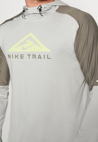 Camiseta de manga larga Nike Trail en gris claro con mangas verde oliva oscuro. Presenta un gran logo triangular amarillo y capucha ajustable.