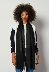 Svart sjal i ett lätt tyg med fransiga kanter, bärs över en vit topp och en mönstrad stickad tröja med mörka ärmar.