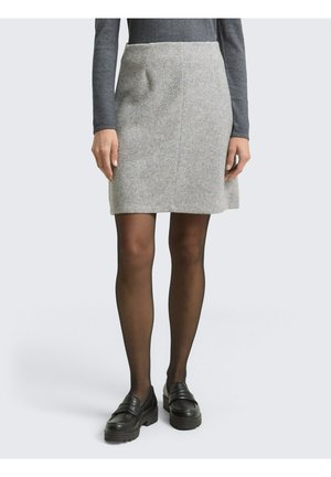 Frau trägt einen grauen Strickrock, dunkle transparente Strumpfhosen, schwarze klobige Loafers und ein langärmeliges dunkelgraues Oberteil, steht vor einem schlichten Hintergrund.