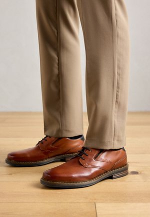 Pantofi din piele maro cu șireturi purtați cu pantaloni bej pe un podea din lemn.