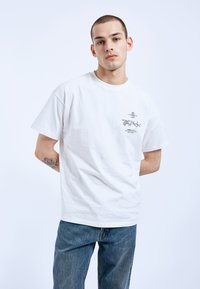 Bílý bavlněný tričko s kulatým výstřihem, krátkými rukávy a grafickým potiskem na levé straně hrudi. V kombinaci s modrými džínami.