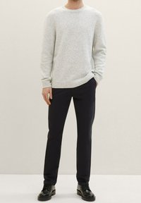 Maglione grigio chiaro lavorato a maglia con scollo rotondo e polsini a costine, abbinato a pantaloni blu scuro e scarpe nere. Tessuto morbido, vestibilità casual.