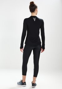 Svart långärmad träningsskjorta med en texturerad halsdetalj, tillsammans med svarta leggings med en vit sidrand. Buren med grå sneakers.