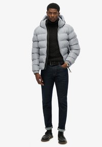 Giacca puffer grigio chiaro con cappuccio, indossata sopra un maglione nero a trecce. Abbinata a jeans in denim scuri e scarpe nere.