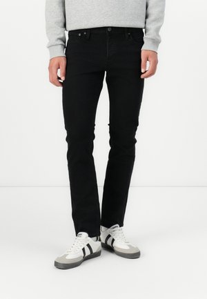 Muž stojící opřený o bílé pozadí, oblečený v úzkých černých džínách, šedé mikině a bílých teniskách s černými pruhy.