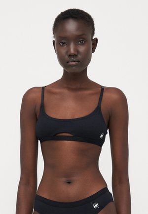 Black ribbed bralette with narrow straps and a front cut-out detail. Features a small logo on the left side and soft, stretchy fabric.