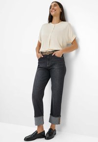Lächelnde Frau, die an einer Wand lehnt, trägt eine cremefarbene Kurzarmbluse, dunkelblaue, hochgekrempelte Jeans mit Leopardenmuster und schwarze Slipper.