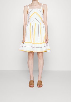 Kvinde iført hvid ærmeløs kjole med gule og brune lodrette og chevronstriber, flæsekant og vævede brune slip-on sko.