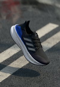 Black running shoe with three silver stripes, blue accents, textured mesh upper, white sole, and "LIGHTBOOST" lettering on the side.