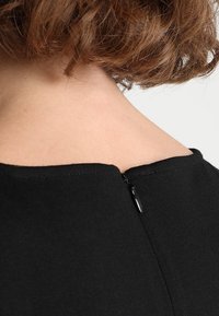 Czarna odzież z zaokrąglonym dekoltem i krótkim suwakiem z tyłu. Gładka tekstura o prostym wzornictwie bez widocznych wzorów.