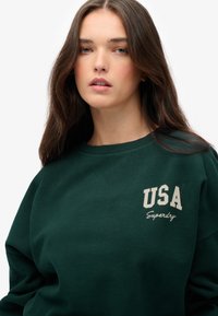 Sudadera verde hecha de un tejido suave con cuello redondo. Presenta 'USA' y 'Superdry' en impresión de color claro en el pecho.