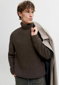Un pull en maille marron avec un col haut, présentant un motif texturé, des poignets côtelés et une coupe ample, porté avec un manteau gris clair drapé sur une épaule.