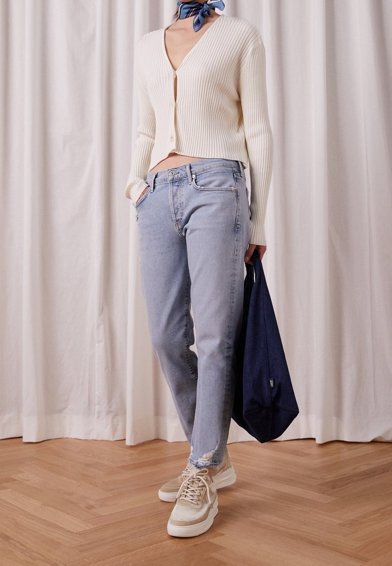 Femme portant un cardigan côtelé crème, un jean bleu clair, des baskets beiges, un foulard bleu autour du cou, tenant un sac cabas bleu marine, debout sur un sol en bois avec un fond de rideau.