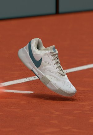 Weißer Nike-Sneaker mit blauem Swoosh und beigen Schnürsenkeln in der Luft über einem roten Sand-Tennisplatz mit weißen Linien.