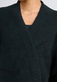 Närbild på en person som bär en svart, texturerad fleecetröja med djup V-ringning över en svart innertröja.